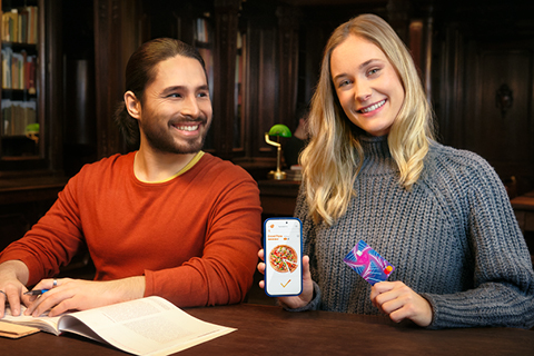Zwei junge Menschen sitzen in einer Bibliothek. Sie bestellt über das Handy eine Pizza und bezahlt rasch und unkompliziert mit ihrer Debit Mastercard.