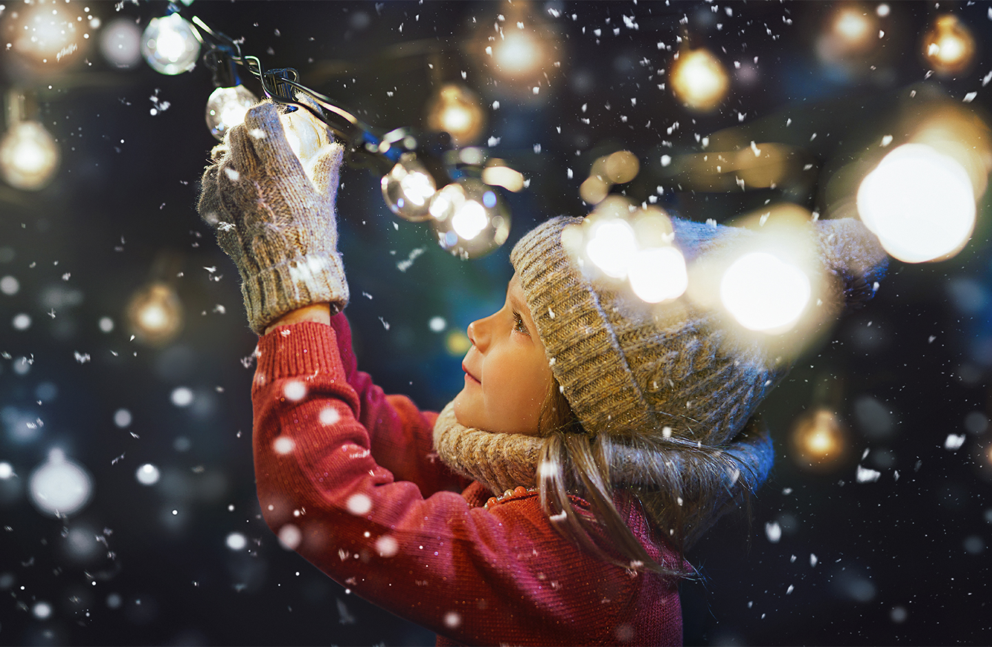 Das Bild zeigt ein winterlich bekleidetes Mädchen nachts bei Schneefall - sie hält ein Licht einer Lichterkette in den Händen.