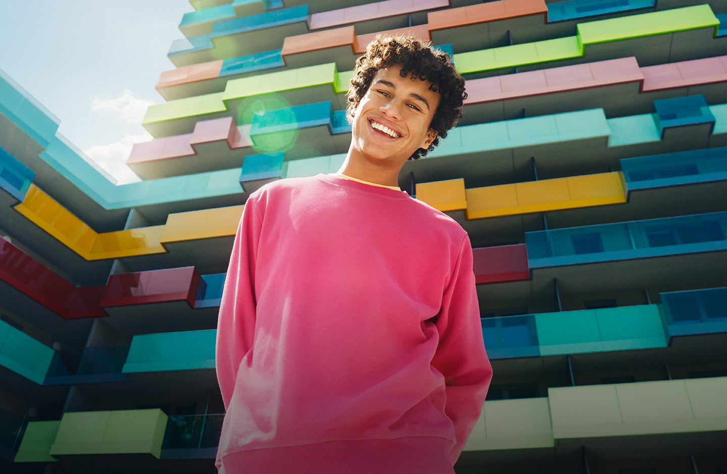 Ein junger Mann in einem rosa Pullover steht vor einem Hochhaus und lacht in die Kamera.