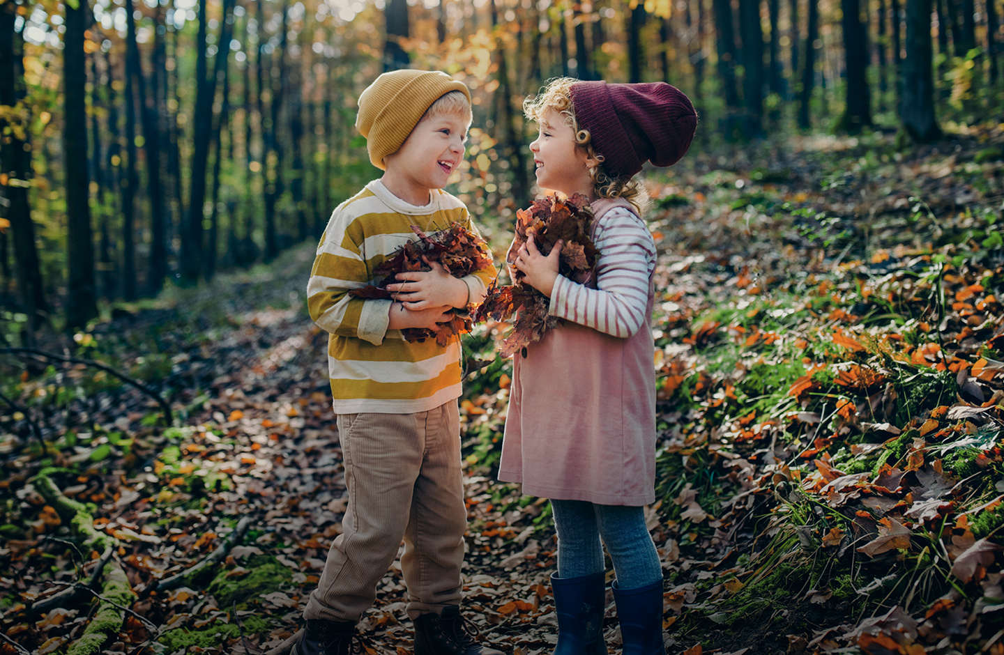 Zwei Kinder stehen in einem herbstlichen Wald. Beide haben trockene Blätter in den Armen.