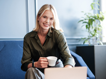 Eine Frau sitzt vor ihrem Laptop auf einem Stuhl mit einer Kaffeetasse in der Hand