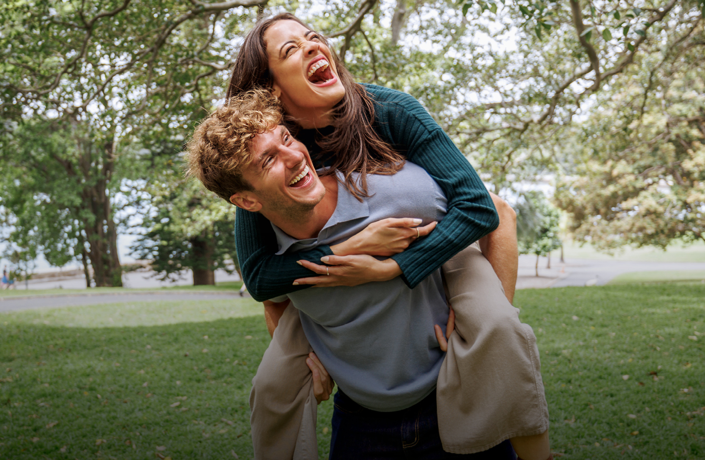 Ein Mann trägt eine Frau Huckepack in einem Park - beide lachen.
