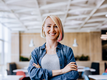 Eine Frau mit kurzen, blonden Haar verschränkt in einem Büro lachend die Arme.