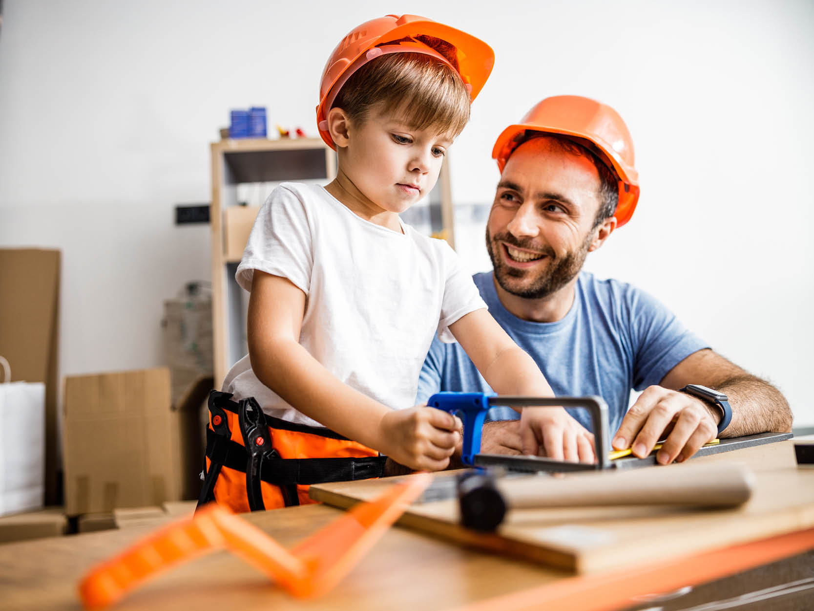 Ein Junge sägt mit seiner Spielzeug-Säge an einem Holzstück, neben ihm lachend sein Vater.