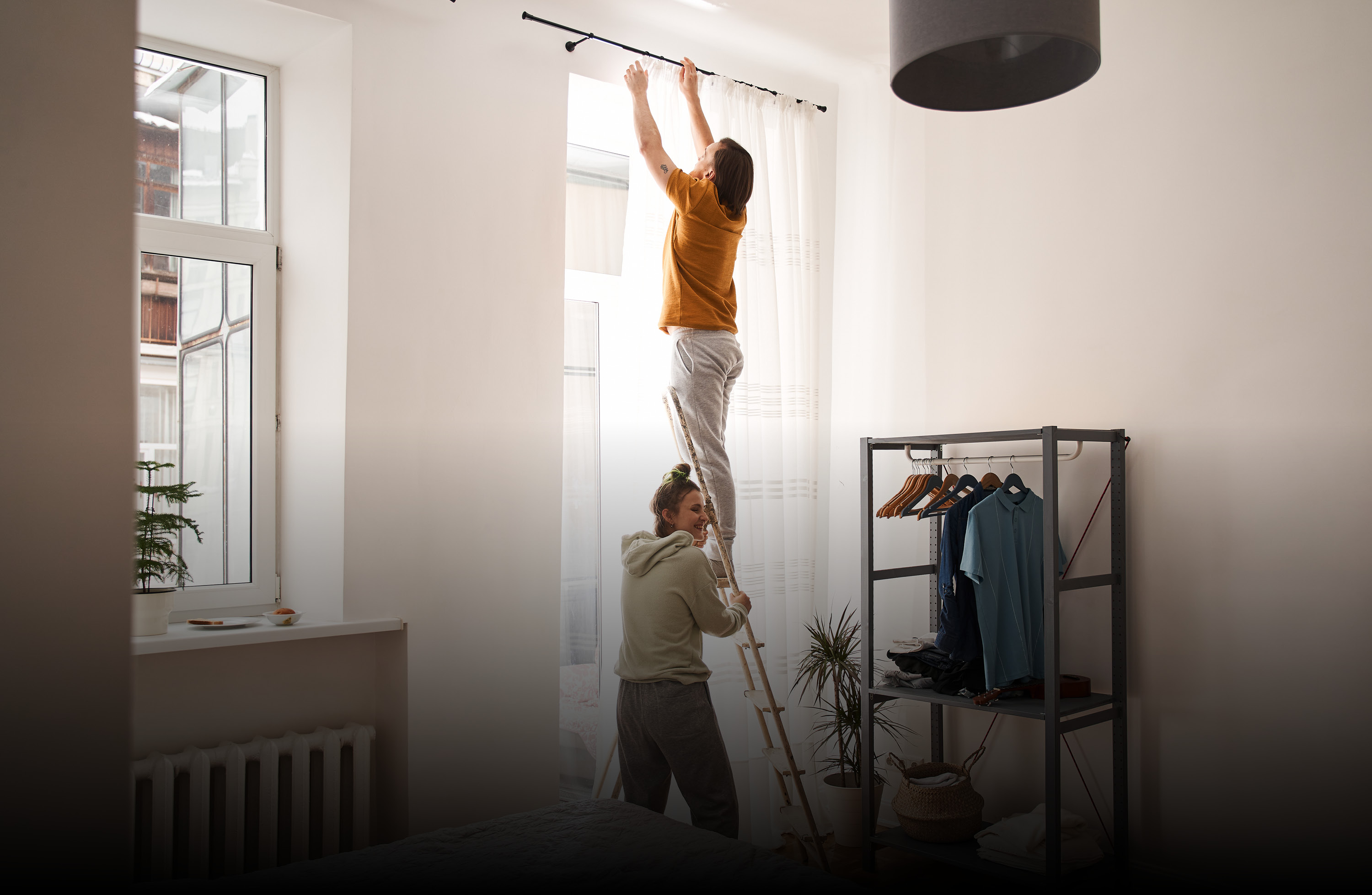 Zwei Personen montieren eine Vorhangstange in einem hellen Schlafzimmer, wobei eine Person auf einer Leiter steht.