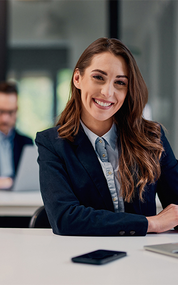 Eine Geschäftsfrau im Blazer sitzt lächelnd an einem Schreibtisch.