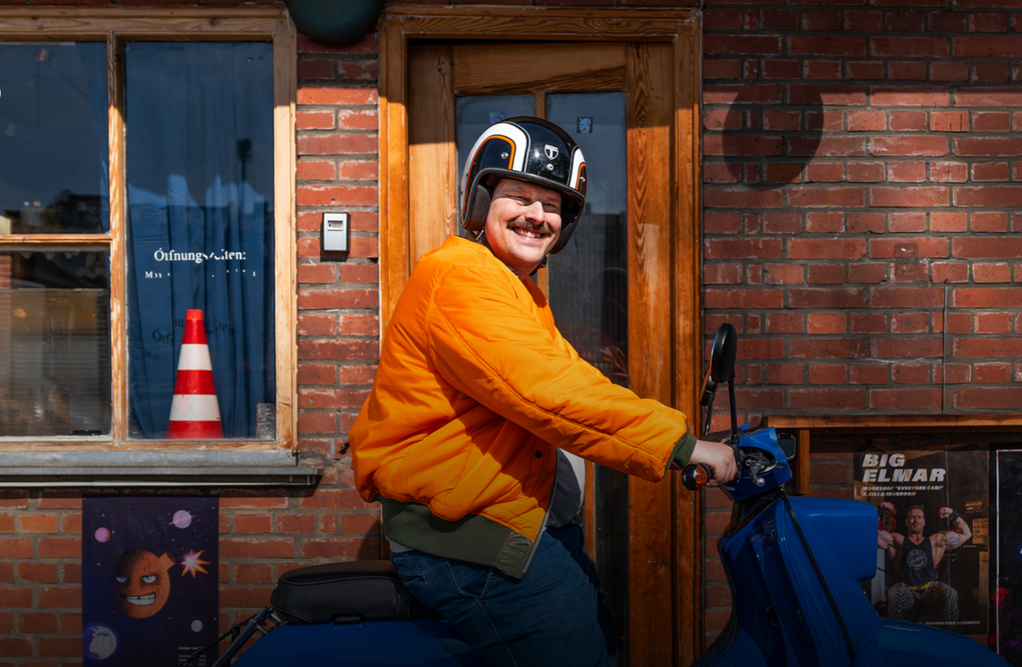 Ein Mann in oranger Jacke und blauem Helm sitzt auf einem Moped und lacht.
