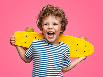 Ein Bub mit gelockten Haaren und einem gelben Skateboard lacht in die Kamera.