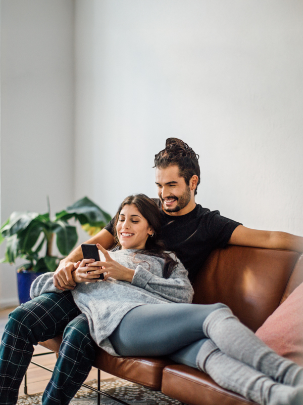 Eine Frau liegt in den Armen eines Mannes auf einer Couch. Gemeinsam blicken sie auf ihr Handy.